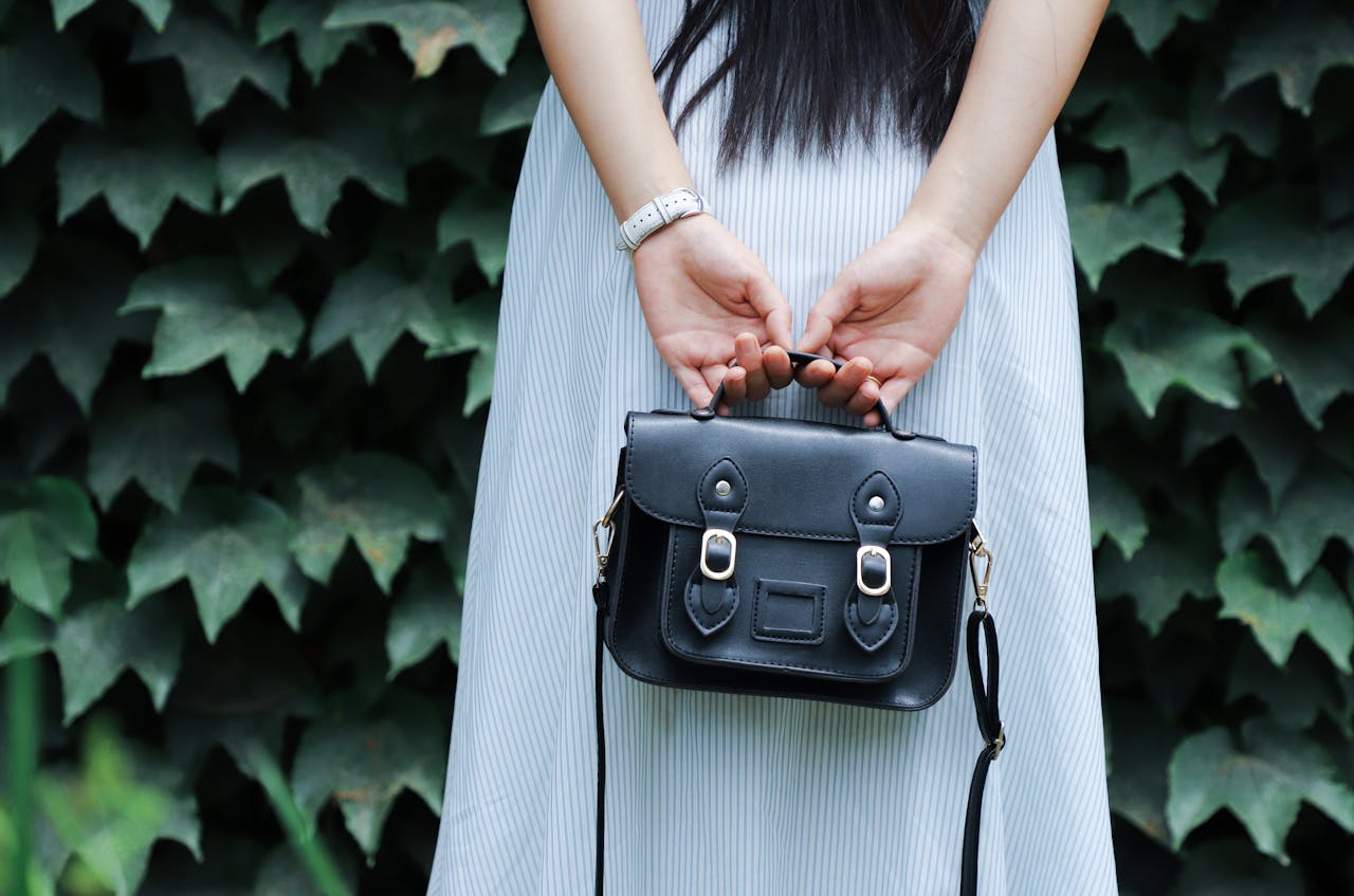 hero-img-02 A woman in a striped dress holds a black leather handbag with lush green leaves in the background.