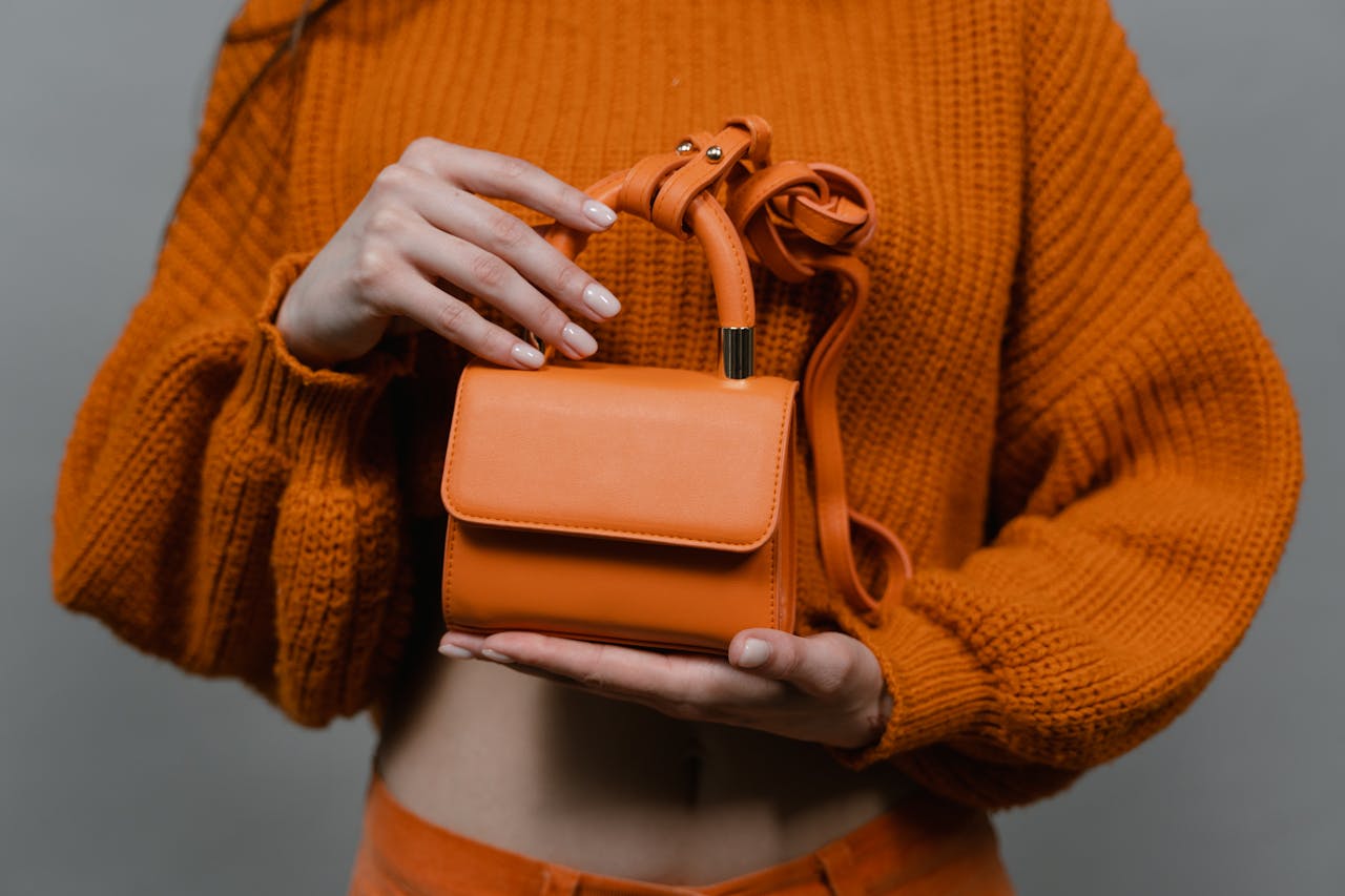 about-01 Woman holding an orange leather mini handbag against an orange sweater in a studio setting.